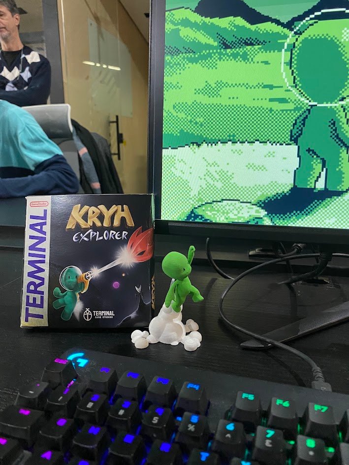 Caixa do jogo Kryh Explorer e um bonequinho alienígena verde em frente a uma tela em pixel art