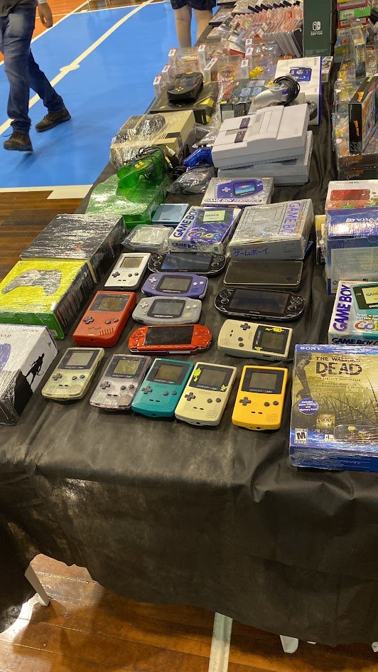 Uma mesa cheia de Game Boys, PSPs e consoles antigos numa feira de games retrô