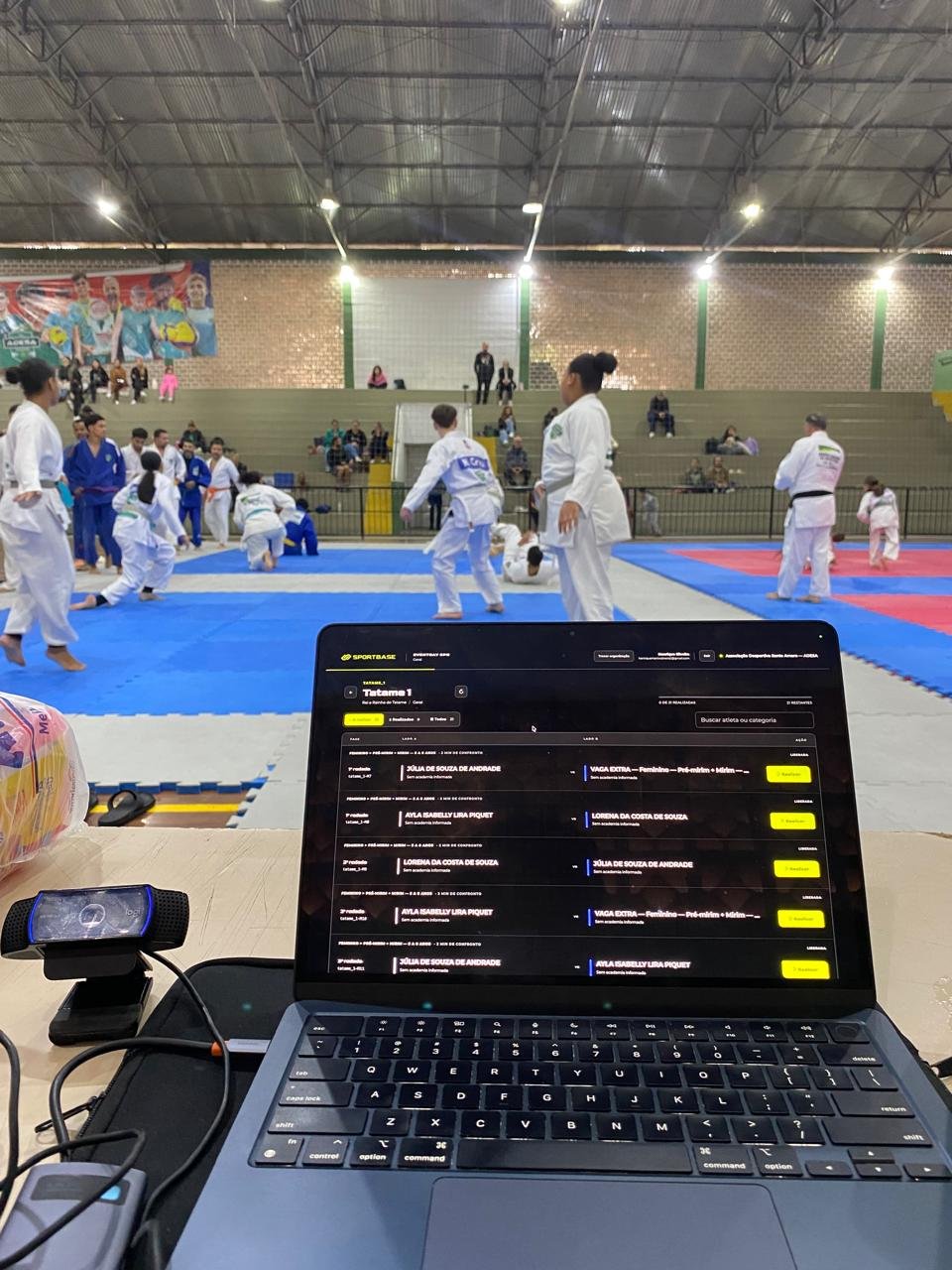 Notebook com a chave do tatame no Sportbase, em frente a um campeonato de judô num ginásio