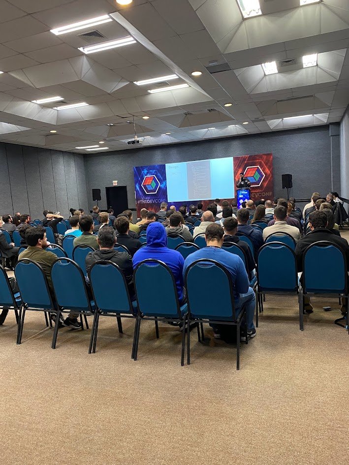 Plateia numa sala de conferência assistindo uma palestra, com o logo do The Developers Conference no telão